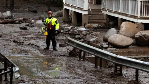 Reuters A male first responder dressed in neon yellow PPE surveys debris and mud covering a roadway in Ruidoso, New Mexico.