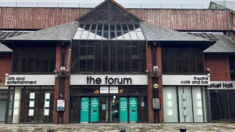 The Forum in Barrow, a large modern building of red brick and some wood cladding at roof level, with a mainly glass entrance foyer and containing a market and theatre. Signs above its glass doors read 'arts and entertainment', 'theatre', 'cafe and bar', and 'market hall'. The building closed in 2024.