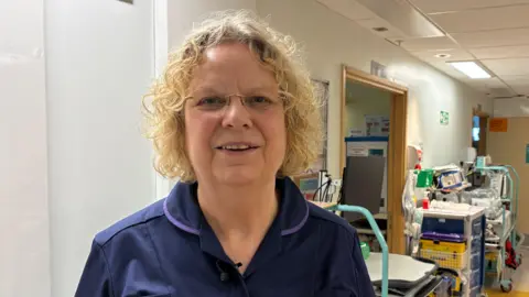 A woman with curly blonde hair smiles at the camera. She is wearing a blue uniform. There is hospital equipment in the background.