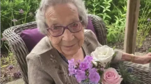 Handout Betty, a woman with short grey hair and purple glasses, sitting in a wicker chair with a purple cushion. She is holding a bunch of pink and white flowers in one hand while grabbing a walking frame out of shot with her other hand. There is greenery in the background. 