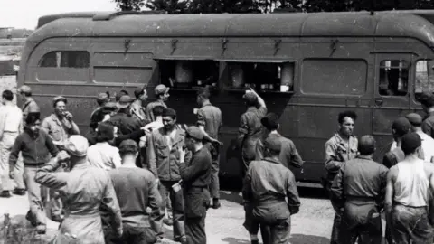 IWM/Roger Freeman Collection USAAF personnel receive food and drink from an American Red Cross Clubmobile. The vehicle is a bus which has two open flaps in its wall. Men are reaching up to the counter behind the window. Others are milling around, chatting and eating