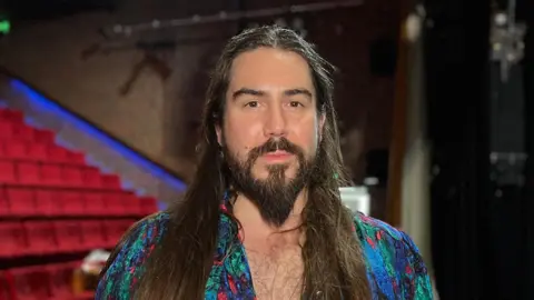 Adam has long brown hair and a beard. He is wearing a multi-coloured top.