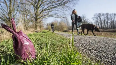 A rural or park‑like setting on a clear day, with a gravel path running diagonally from the background toward the foreground. In the immediate foreground, a small pink or purple tied dog poo bag sits on the grass beside the path.
In the middle ground, a person is walking along the gravel path while holding a leash attached to a dog. The dog is positioned slightly ahead and to the side of the person, with its nose close to the ground as if sniffing the grass near the edge of the path. Behind them, another person is visible further down the path, walking in the same direction.
