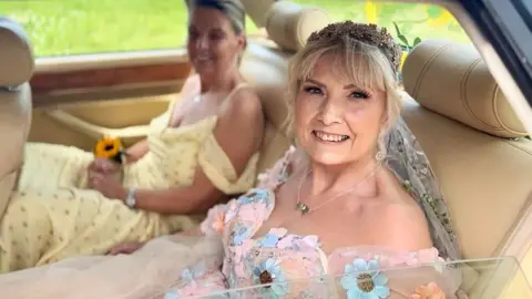 Trasi smiles for the camera. She is the back seat of a care looking out the window. She is wearing a wedding dress, a shoulder cut dress with pink sheer and blue and orange flowers sewn on. there is a woman in the seat beside her out of focus and wearing a yellow dress. 