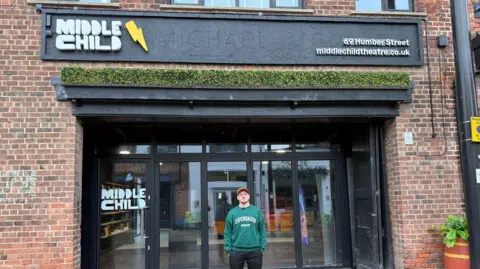 A fully body shot of Paul Smith stood in front of Middle Child theatre with his hands in his pocket, looking at the camera. He is wearing glasses, an orange cap, a green sweater with "Copenhagen" in white bold capital letters printed on the front and black trousers.