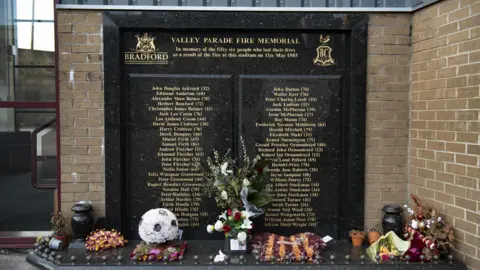 Getty Images A stone memorial with floral tributes laid at the foot of it. A list of 56 names, 28 on either side, are engraved on it, with their ages next to them.