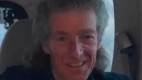 Alban Watts, who has curly grey hair with some of it swept back, smiles at the camera. He is sat in a white seat and is wearing a navy shirt.