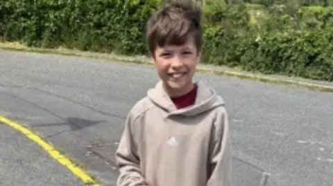 Destin has brown hair and is wearing a pale brown hoodie in this picture. he is standing besides the road and smiling.