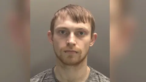 A police-issued mugshot of Kieran Williams. He has a neutral expression, fair short hair and a double bottom lip piercing. He is wearing a grey t-shirt.