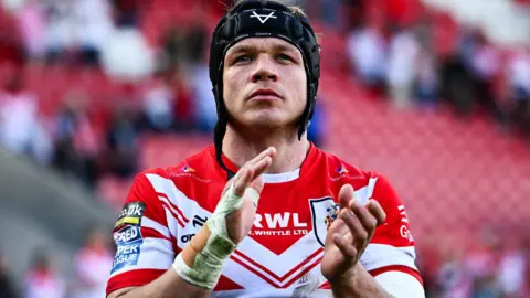 Jonny Lomax, wearing a trademark scrum cap with the chin strap undone, applauds the St Helens fans after a match