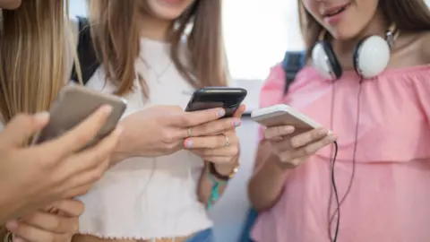 Getty Images Three teenage girls huddled around using mobile phones