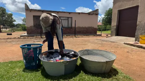 A woman bends over two large pans of murky water, one containing laundry, on a small area of grass.