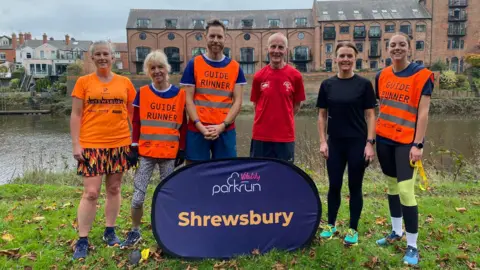 Shropshire Shufflers Sam, along with two women to his left and a man and two women to his right, are standing in front of the purple Shrewsbury parkrun banner, with the river Severn in the background. Sam and two of the women are wearing bright orange vests that say "guide runner".