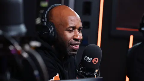 Sarah Janes/BBC DJ Spoony speaking into a large microphone with the BBC Radio 2 logo. He is wearing headphones and is smiling.