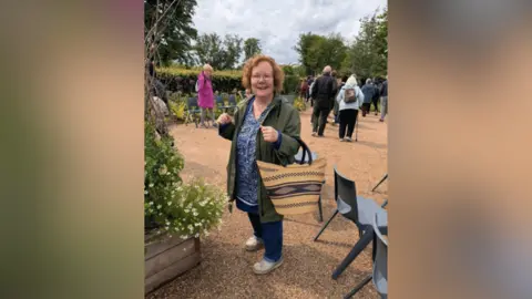 Alison Butt Alison Butt stands in an outdoor garden centre smiling. She has a woven basket hanging from her arm. 