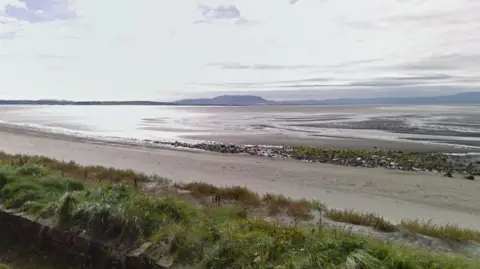 A sandy beach at low tide, with the beach going into the water in the distance. Hills are visible in the background. It is a sunny day, with sparse clouds in the sky.