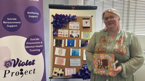 There is a purple and white upright banner with slogans about the violet project on the left, in the middle there is a cork board with some postcards on and on the right is the founder of the charity, Mel Griffin, who has blonde hair and is wearing glasses and a long light green dress. She is holding a memory book dedicated to her younger sister. 