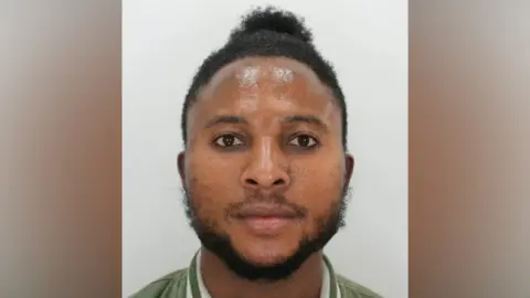 A police custody image of a man. He has short black hair, a black beard and is wearing a green jacket with white stripes around the collar.