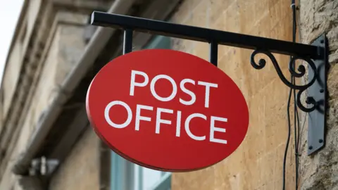 Getty Images A post office sign is hanging off the side of a stone wall.