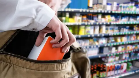 Getty Images A white hand places a box in a bag secretly, with grocery shop shelves blurred in the background