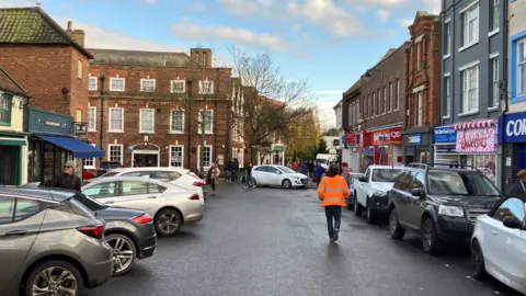 Guy Campbell/BBC Cars parked in a town centre street close to shops. A person wearing a fluorescent orange jacket is walking in the road