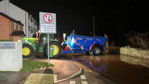 A green tractor has a blue tanker attached to its rear. They are sat across a large area of a flooded road. A stone wall is behind the blue tanker.