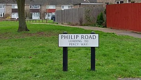 An estate made up of light brown terrace houses. In the foreground on a patch of grass is a sign which says Philip Road, leading to Percy Way