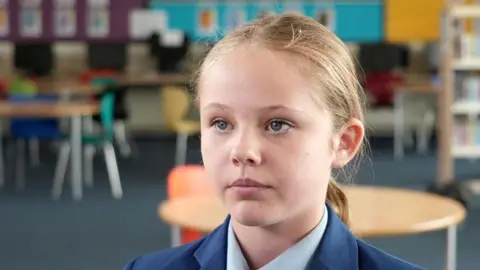 Shaun Whitmore/BBC A head and shoulders image of a young woman. She is wearing a navy blazer over a smart shirt. She is looking away from the camera. She is sitting in a classroom and there are chairs and tables behind her.