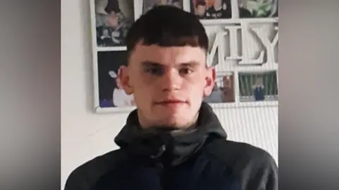 Kieran Williams who has short, brown hair and a bit of stubble on his chin. He is wearing a grey hoodie which is zipped all the way to the top. He is standing in front of a collection of photos which says 'family' in the middle. The photo is slightly pixilated, like someone has taken a photo of the picture on another screen.