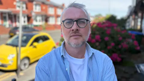 Billy is stood outside his house and looking at the camera. He has a white t-shirt on under a blue shirt and is wearing glasses. A yellow car can be seen behind as we all a floral bush to his right.