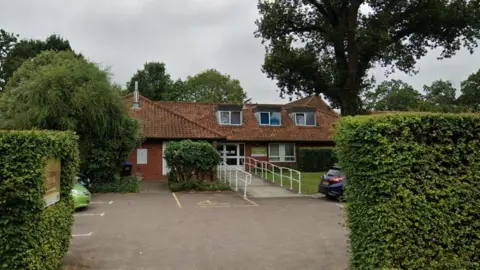 Google Exterior of a single-storey brick care home with a tiled roof, a ramp leading to the entrance, and a small car park bordered by hedges and trees.