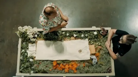 Overhead shot, showing a human composting vessel filled with flowers and organic matter, with a small white sheet laid on the top. In front of the vessel is Kristopher Hughes, wearing a colourful shirt, and opposite at the end of the vessel is one of Return Home's staff members, a women dressed in a black t-shirt with her hair tied back