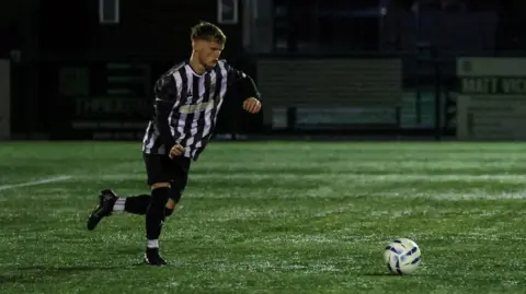 Karen Harland Photography Kory Whitfield is on a football pitch running towards a football. He is wearing a black and white football kit.