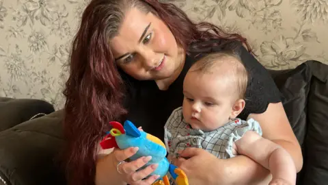 a woman sitting on her sofa with her baby on her lap playing with a soft toy together