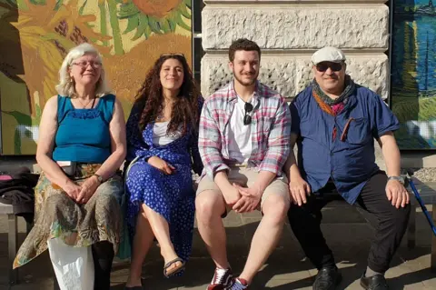 Supplied Two women and two men sit on a bench in the sunshine outside a building with two murals on panels of artwork by Van Gogh.