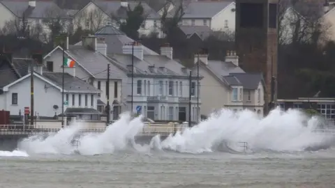 Getty Images Waves hitting the sides of a coastal route with multiple buildings behind.