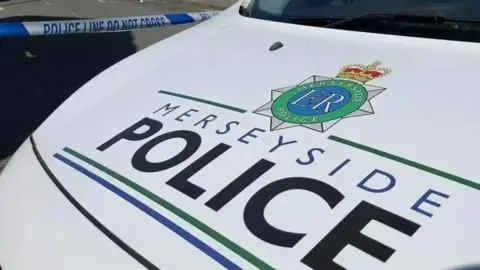 The white bonnet of a Merseyside Police patrol car, bearing the name of the force in black and blue lettering. Police tape can be seen across a road behind the vehicle.