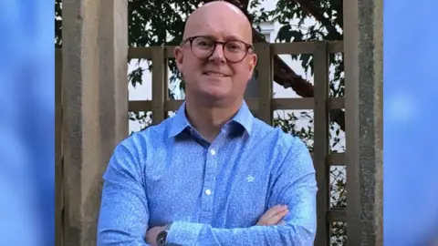 Supplied Tim Wickens is smiling with his arms crossed in front of a stone fence. He is wearing a blue shirt and a watch. He has black-rimmed round glasses.