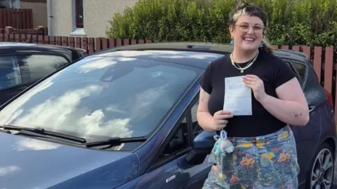 Lois Stevenson A woman standing in front of a blue car holding a document, with a residential street and wooden fence in the background.