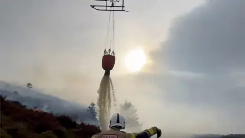 Picture of a water bucket dropping a lashing of water onto the mountainside. A firefighter can be seen with their back to the camera, they wear a white helmet and a fire coat. Smoke can be seen rising from the trees on the mountainside. The sun beams in the background. 