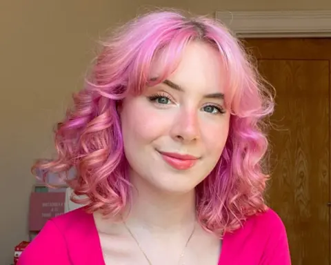 Milly Evans A woman with wavy pink hair in a pink top smiles at the camera for a selfie, taken indoors