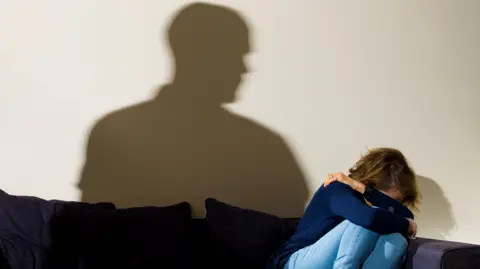 Woman sitting on a navy sofa cowering with her knees to her chest and covering her face with her arms. 