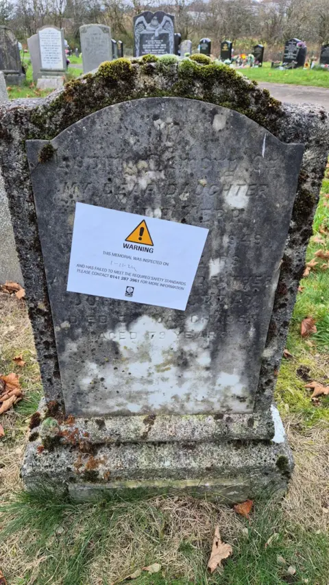 A gravestone in Riddrie Park Cemetery. It is very worn, and has a warning sticker applied to it. 