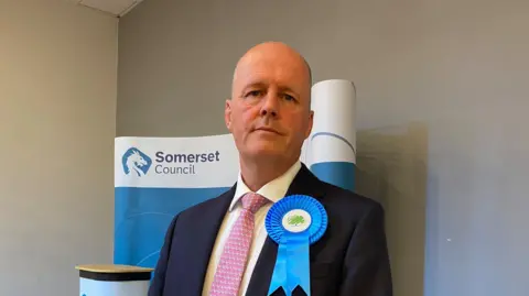 Ashley Fox wearing a white shirt, red patterned tie and black blazer. He has a blue rosette pinned to his left lapel, and is standing in front of a blue Somerset Council banner.