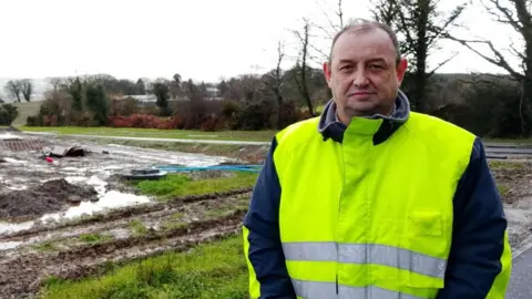 Councillor Declan Norris is standing in a high-vis vest. He is near a marsh ground during the early stages of work on the cemetery.