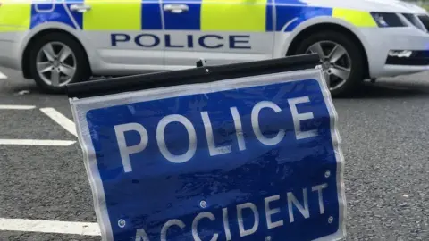 BBC A blue police accident sign resting on the road in the foreground. In the background there is a parked police car with blue and yellow hi-vis decals.