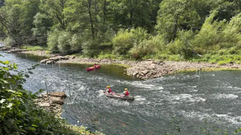 BBC A river with trees near it in the top half of the photo. Two canoes, one grey and one red, are on the river in the bottom half of the image.