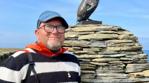 Rob Flett/BBC Jonathan Ford is dressed in a black and white jumper with a badge of a Great Auk on it and is standing next to the statue of the bird.