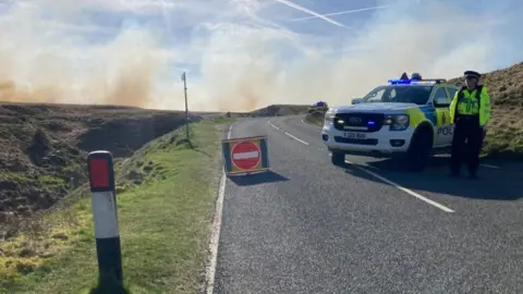 Charles Heslett/BBC A road on a moor which has been blocked off by a police car, with a stop sign put up on the road and a police officer standing nearby. The sky is blue but smoke can be seen rising from the moor. 