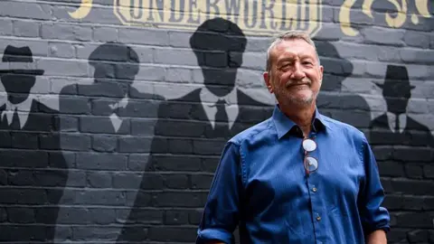 Steven Knight, creator of Peaky Blinders stands with the background of painted silhouettes of his most famous creations behind him at the opening of Peaky Blinders underworld. He is wearing a blue shirt and is smiling